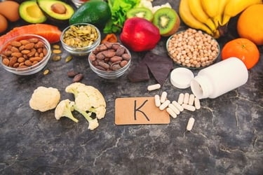 Variety of Vitamin K food sources including leafy greens, nuts, and fruit alongside supplement capsules.