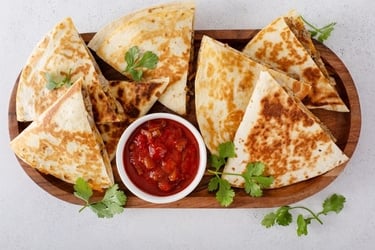 A wooden platter filled with toasted cheese and meat quesadilla triangles, served with fresh salsa and cilantro.
