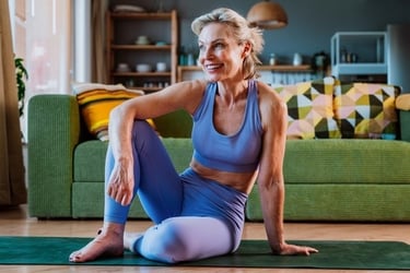 Smiling senior woman in activewear sitting on a yoga mat in her bright living room, enjoying a post-workout moment.