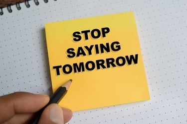 Hand pointing to a yellow sticky note with the motivational quote Stop Saying Tomorrow.