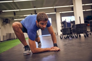 A man in a blue t-shirt performing a deep lizard stretch on a gym floor for mobility training.