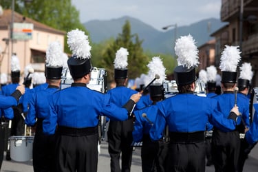 A marching band in blue uniforms with white plumes performs during a parade in a scenic mountain town.