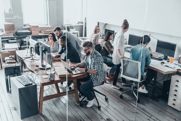 A diverse team of creative professionals working on laptops and computers in a modern open-plan office space.