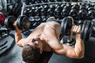 A shirtless man performing a dumbbell chest press on a weight bench in a gym setting.