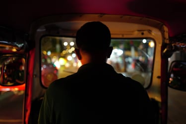 Silhouette of a tuk‑tuk driver in Phnom Penh facing blurred city lights at night, By ACAT Photos.