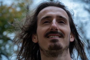 Smiling man with long brown hair and a goatee posing outdoors with wind blowing through his hair.