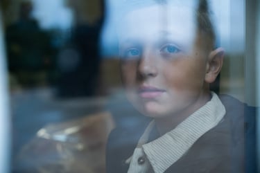 A young boy looking through a glass window with a pensive expression.