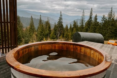 Rustic wood-fired hot tub on a deck with a scenic mountain and forest view under a cloudy sky.