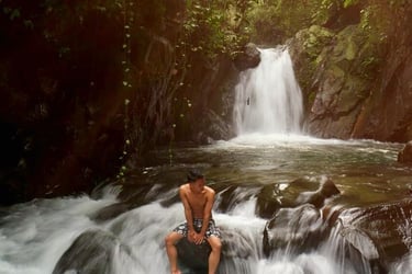 Pria bertelanjang dada duduk di atas bebatuan besar di tengah aliran air terjun deras dengan latar belakang air terjun yang l