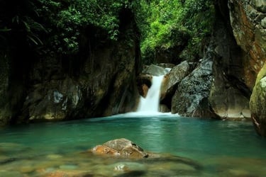 Kolam air terjun alami dengan air berwarna hijau toska yang jernih, dikelilingi tebing batu gelap dan vegetasi hutan hijau le