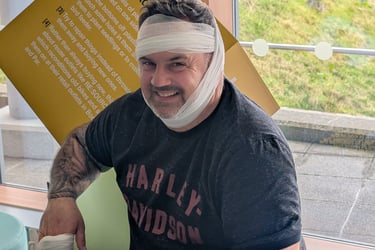 Smiling man with head bandage and hand dressing sitting in a hospital waiting room.