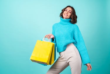 Mujer sonriente con un suéter azul, sosteniendo bolsas de compras amarillas, sobre un fondo turquesa