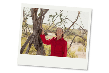 Smiling senior woman in a vibrant red shirt posing outdoors by a desert tree.