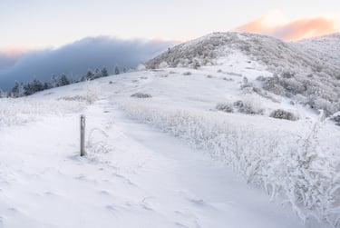 Snow-covered hiking trail leading up a frozen mountain ridge at sunrise with a cloudy sky.
