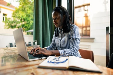 A focused black woman with braids working on a laptop at a cafe table with a notebook.