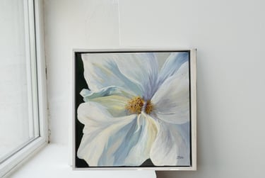 A square  painting of a white flower in a white frame on a windowsill.