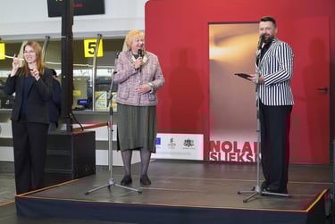 Sieviete runā mikrofonā uz skatuves pie instalācijas Nolaid slieksni, blakus vīrietis un sieviete 