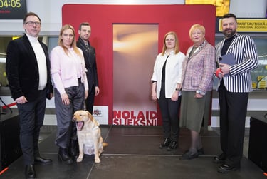 Seši cilvēki stāv pie instalācijas ar uzrakstu “Nolaid slieksni”, priekšplānā sēž suns-pavadonis