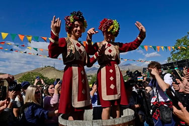 Women in traditional Armenian dress wearing grape headpieces crush grapes at a harvest festival.