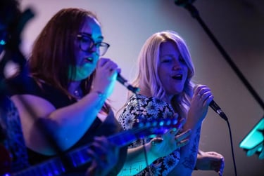 Two female vocalists performing live music on stage with microphones and blue stage lighting.