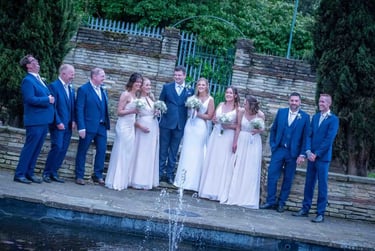 A happy wedding party in blue suits and white bridesmaid dresses posing by a garden fountain.