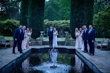 Bride and groom with bridal party posing around a garden fountain at a wedding venue.