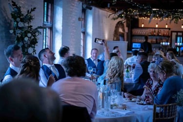 Wedding guests smiling and taking photos with smartphones at a rustic indoor reception dinner.