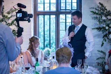 Groom giving a wedding speech at the head table being filmed by a professional videographer.