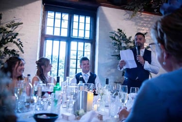 A best man gives a wedding speech at a reception dinner with the smiling bride and groom.