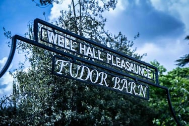 Black wrought iron sign for Well Hall Pleasaunce and Tudor Barn against a blue sky with trees.