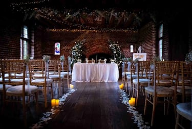 Indoor rustic wedding ceremony venue with brick walls, wooden chairs, floral arches, and candlelit aisle.
