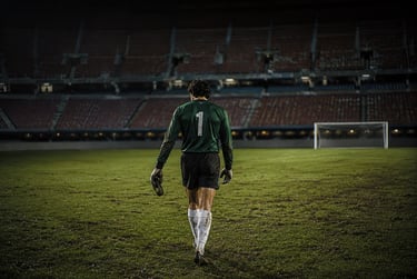 A soccer goalkeeper in a green jersey walks across a dark stadium field toward the goal.