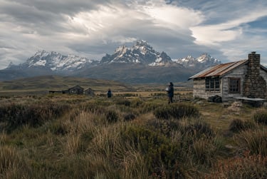 Rustic wooden cabin in the Patagonian wilderness with snowy mountain peaks and dramatic clouds.