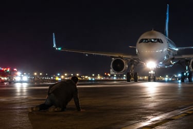 A person kneeling on a dark airport runway at night facing a large commercial airplane with emergency vehicles nearby.