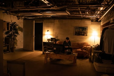 A man sits on a couch in a dimly lit basement living room with exposed ceiling pipes and warm lamp lighting.