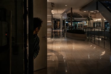 A man peers from a dark doorway into a brightly lit, empty modern shopping mall at night.