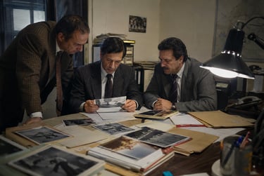 Three male detectives in suits examine crime scene photos on a desk in a dark office.