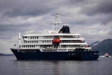 Luxury expedition cruise ship sailing on calm waters near mountains under a cloudy sky.