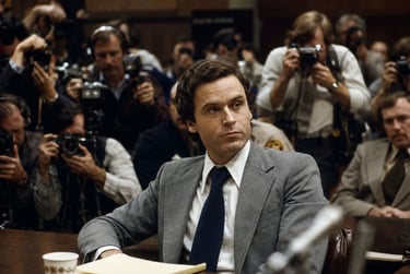 Ted Bundy in a grey suit sits in a crowded courtroom surrounded by photographers and media during his trial.