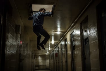 A man in a blue shirt climbs into a ceiling hatch in a dark, narrow prison hallway.