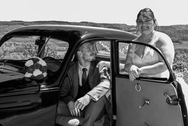 Couple de mariés en noir et blanc posant avec une voiture de collection