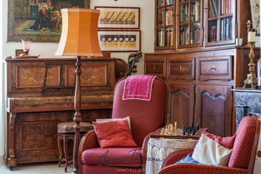 Salon de style vintage avec piano ancien en bois, fauteuils rouges et jeu d’échecs.