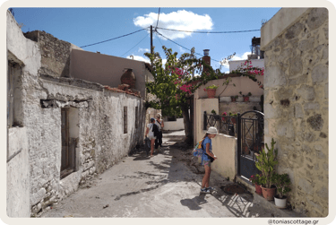 People walking down the streets of Alagni Village, passing stone houses and vibrant summer flowers