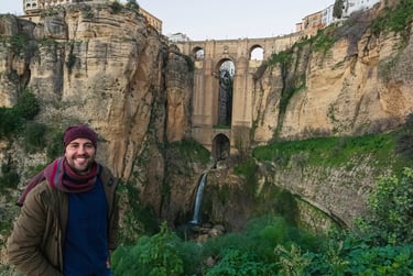 A man smiling in front of the historic Puente Nuevo bridge and waterfall in Ronda, Spain.