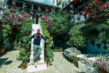 A man posing on white stairs in a traditional Cordoba patio filled with vibrant blooming flowers.