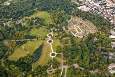 Aerial view of a sprawling city park with lush green trees, circular pathways, and an outdoor amphitheater.
