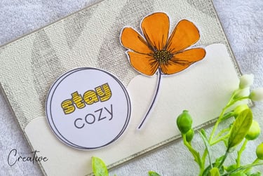 Handmade greeting card with "Stay cozy" sentiment and big orange flower on a beige background.