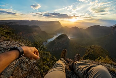 Hiker sitting on a rocky cliff edge overlooking a scenic mountain valley and river at sunrise.