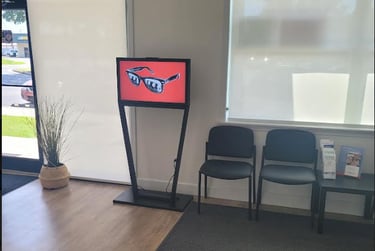 A digital signage display showing eyewear in an optometry office waiting room with black chairs.