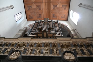 Low angle view of a historic pipe organ and ornate stone carvings under a geometric wooden church ceiling.